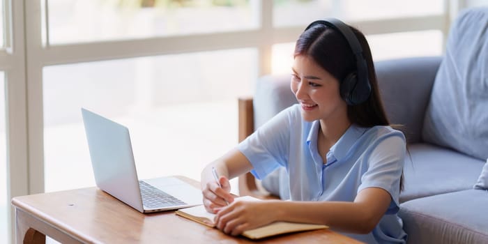 Online education, e-learning. Asian woman in stylish casual clothes, studying using a laptop, listening to online lecture, taking notes, online study at home.