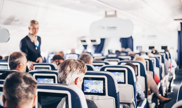 Interior of airplane with passengers on seats and stewardess in uniform walking the aisle, serving people. Commercial economy flight service concept