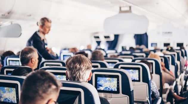 Interior of airplane with passengers on seats and stewardess in uniform walking the aisle, serving people. Commercial economy flight service concept