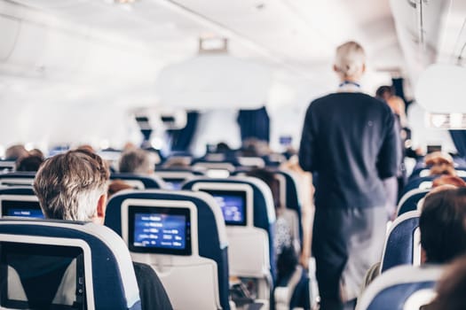 Interior of airplane with passengers on seats and stewardess in uniform walking the aisle, serving people. Commercial economy flight service concept