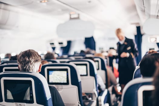 Interior of airplane with passengers on seats and stewardess in uniform walking the aisle, serving people. Commercial economy flight service concept