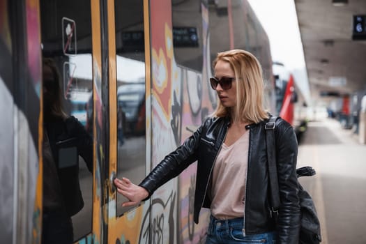 Young blond woman in jeans, shirt and leather jacket wearing bag and sunglass, presses door button of modern speed train to board on train station platform. Travel and transportation.