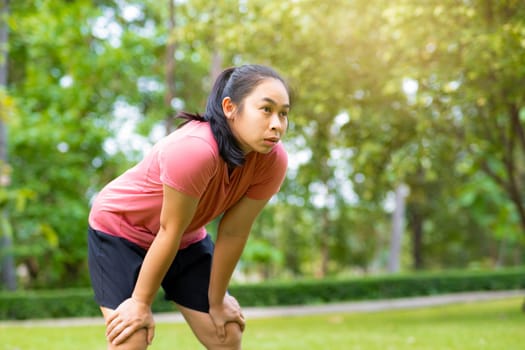 Young Asian woman standing bent over and breathing after running in the park. Young healthy woman taking a break after running. Healthy lifestyle concept.