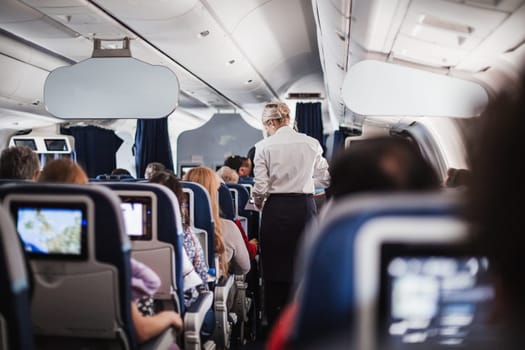 Interior of airplane with passengers on seats and stewardess in uniform walking the aisle, serving people. Commercial economy flight service concept