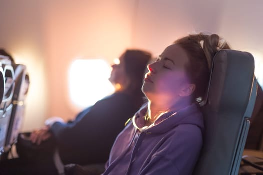 Out of fokus image of tired blonde casual caucasian woman wearing sporty hoodie napping on seat while traveling by airplane. Commercial transportation by planes. Authentic image of real people