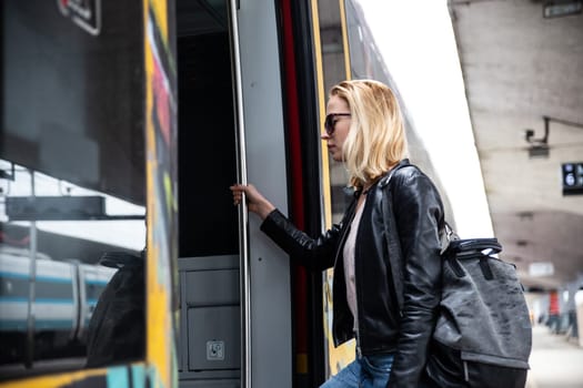 Young blond woman in jeans, shirt and leather jacket wearing bag and sunglass, presses door button of modern speed train to board on train station platform. Travel and transportation.