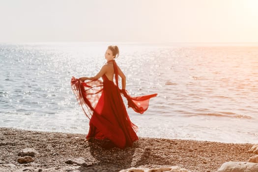 Side view a Young beautiful sensual woman in a red long dress posing on a rock high above the sea during sunrise. Girl on the nature on blue sky background. Fashion photo.