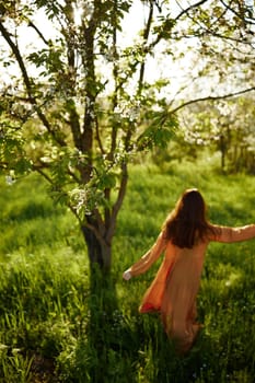 a beautiful, joyful woman is standing in a long orange dress, in the countryside, near a tree blooming with white flowers, during sunset, illuminated from the back and joyfully spinning. High quality photo