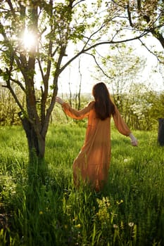 a beautiful, joyful woman stands in a long orange dress, in the countryside, near a tree blooming with white flowers, during sunset, illuminated from the back and reaching for the branches of the tree. High quality photo