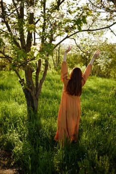 a beautiful, joyful woman stands in a long orange dress, in the countryside, near a tree blooming with white flowers, during sunset, illuminated from behind and raises her hands up while standing with her back to the camera. High quality photo