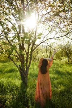 a beautiful, joyful woman stands in a long orange dress, in the countryside, near a tree blooming with white flowers, during sunset, illuminated from behind and raises her hands up while standing with her back to the camera. High quality photo