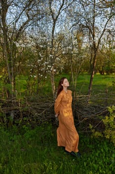 an elegant, sophisticated woman poses relaxed standing near a wicker fence at the dacha in a long orange dress enjoying the silence and peace. High quality photo