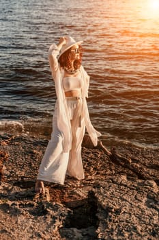 woman sea sunset. woman in a white pantsuit and hat is standing on the beach enjoying the sea. Happy summer holidays.