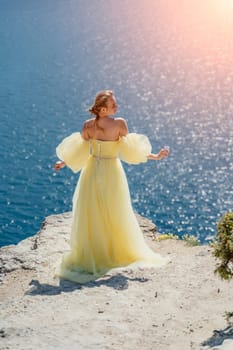 Woman yellow dress sea. Side view Young beautiful sensual woman in yellow long dress posing on a rock high above the sea at sunset. Girl in nature against the blue sky.