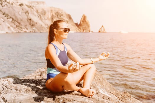 Yoga on the beach. A happy woman meditating in a yoga pose on the beach, surrounded by the ocean and rock mountains, promoting a healthy lifestyle outdoors in nature, and inspiring fitness concept