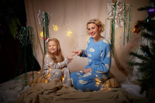 Cute mother and daughter in pajamas having fun in the room with Christmas garlands and white background. The tradition of decorating the house for holidays. Happy childhood and motherhood