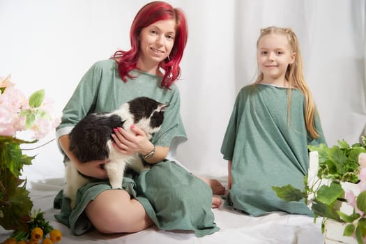 Amazing pretty mother and daughter having fun with flowers and cat in 8 March or in Mother's day. Red haired mom and small little blonde girl having lovely time on white background in studio with pet