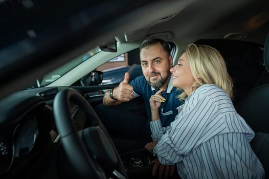 Happy caucasian couple is sitting in a new car in a car dealership. Man showing thumbs up