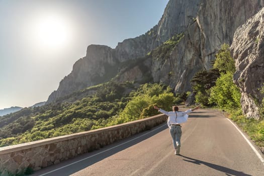 A woman runs along an asphalt road in the middle of a mountainous area. She is dressed in jeans and a white shirt, her hair is braided. A traveler in the mountains rejoices in nature