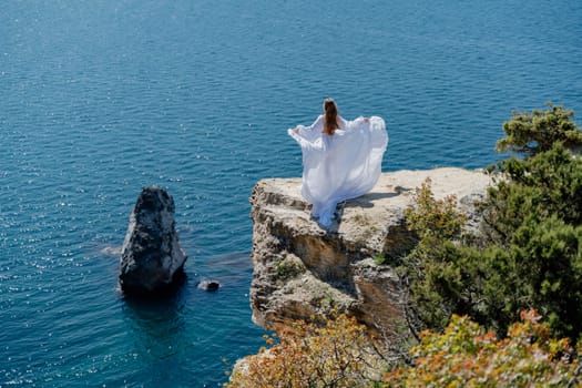 Woman in a white dress on the sea. Side view Young beautiful sensual woman in white long dress posing on a rock high above the sea at sunset. Girl in nature against the blue sky.