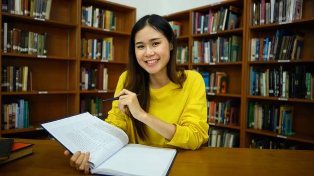 Smiling female student sitting among bookshelves in library reading books for studying and research. Education concept.