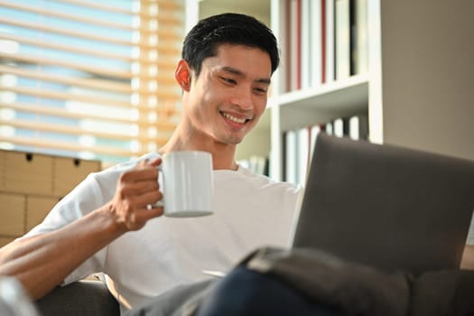 Pleasant man freelancer drinking hot coffee and checking news or email on laptop. People, technology and lifestyle concept.