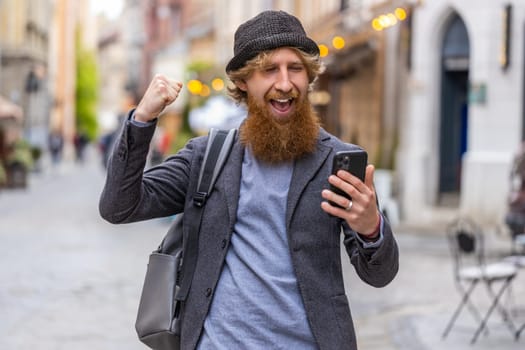 Young bearded man use mobile smartphone celebrating win good message news, lottery jackpot victory, giveaway online. Happy redhead guy walking outdoors in urban city street background. Lifestyles