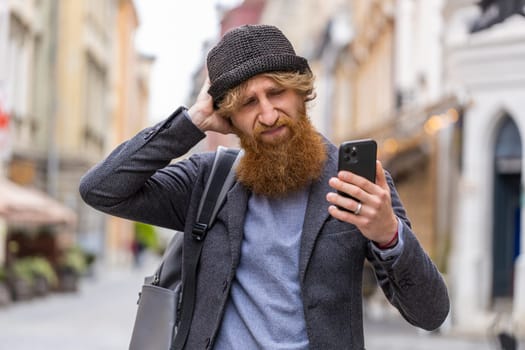 Young bearded man use smartphone typing browsing, loses becoming surprised sudden lottery results, bad news, fortune loss, fail. Redhead guy walking in urban city street outdoors. Town lifestyles