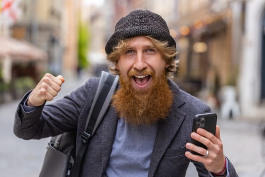 Young red hair bearded man use mobile smartphone celebrating win good message news, lottery jackpot victory, giveaway online. Happy redhead guy walking outdoors in urban city street. Town lifestyles