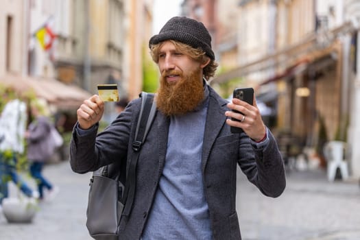 Young bearded man using credit bank card smartphone while transferring money, purchases online shopping, order food delivery, booking hotel room. Redhead guy walking in urban city street outdoors