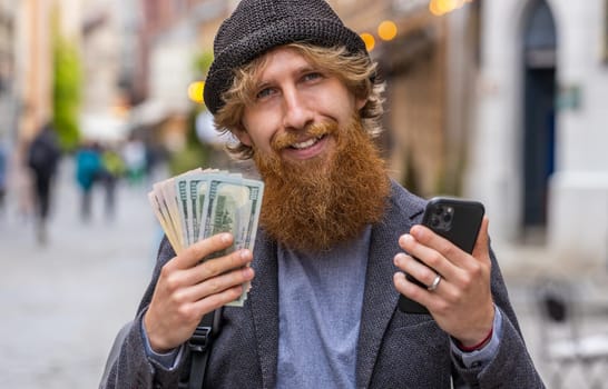 Happy young bearded man counting money dollar cash, use smartphone calculator app, satisfied of income for planned vacation gifts. Redhead guy walking in urban city street. Town lifestyles outdoors