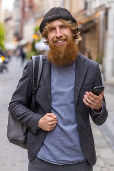 Smiling young man using smartphone typing text messages browsing internet, finishing work, looking at camera. Redhead bearded guy walking in urban city street. Town lifestyles outdoors. Vertical
