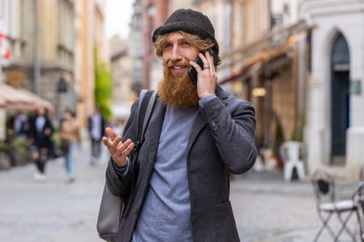 Happy bearded man having remote conversation communicate speaking by smartphone. Handsome young adult guy talking on phone unexpected good news gossip walking in city street. Town lifestyles outdoors