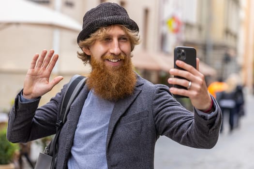 Young bearded man blogger taking selfie on smartphone, communicating video call online with subscribers, recording stories for social media vlog outdoors. Redhead guy walking in urban city street