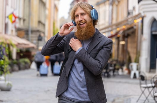 Happy relaxed overjoyed young adult man in wireless headphones choosing, listening favorite energetic disco rock n roll music in smartphone dancing outdoors. Bearded guy walking in urban city street
