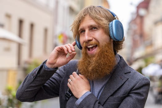 Happy relaxed young stylish rocker man in wireless headphones choosing, listening favorite energetic disco rock n roll music in smartphone dancing outdoors. Joyful guy walking in urban city street