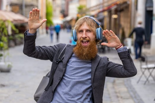 Happy overjoyed young man in wireless headphones listening favorite energetic music celebrating win. Redhead bearded guy tourist traveler walking in urban city street. Town lifestyles outdoors weekend