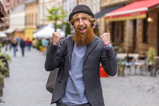 Portrait of young man shouting, celebrating success, winning, goal achievement, good news, lottery jackpot luck, victory outdoors. Redhead bearded guy walking in urban city street. Town lifestyles