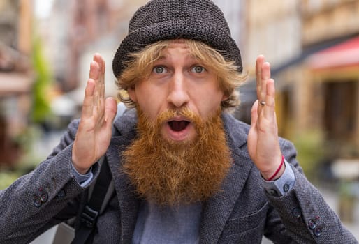 Oh my God, Wow. Young bearded man looking surprised at camera with big open eyes, shocked by sudden victory, game winning, lottery goal achievement, good news outdoors. Redhead guy in city street