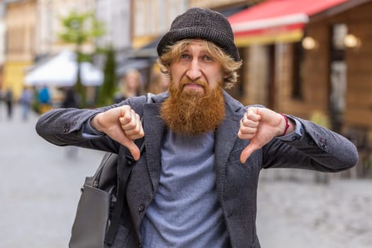 Dislike. Upset young bearded man showing thumbs down sign gesture, expressing discontent, disapproval, dissatisfied bad work, mistake. Displeased redhead guy walking in urban city street outdoors