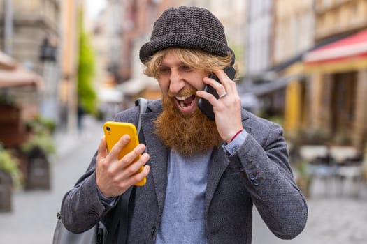Irritated nervous stressed business man talking screaming on two mobile phones having conversation conflict quarrel complaint dispute discuss solve problem outdoors. Guy walking in urban city street