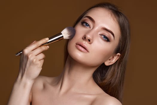 young woman applying blush on cheeks with make-up brush on beige background