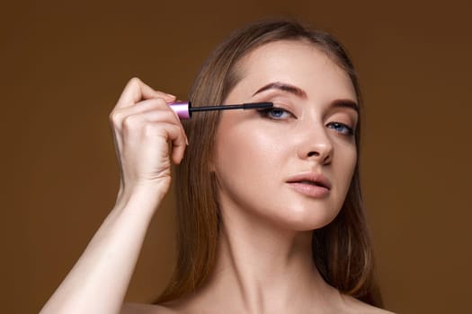 Beautiful woman applying black mascara on long thick eyelashes with brush on beige background
