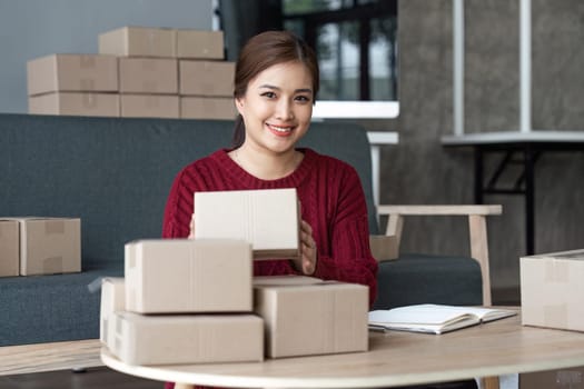 Happy young asian woman startup small business freelance holding parcel box and computer laptop and sitting sofa, Online marketing packing box delivery concept.