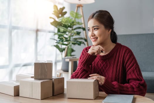 Young asian woman startup small business freelance sitting with parcel box and computer laptop on table in living room at home, Online marketing packing box delivery concept.