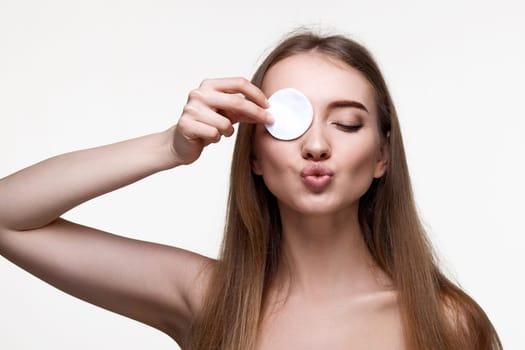 beautiful positive woman with naked shoulders close one eye with white cotton pad isolated on white background
