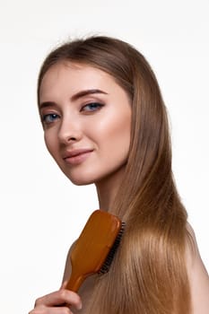 Beautiful smiling brunette woman looks at the camera and combs her hair with wooden brush isolated on white background