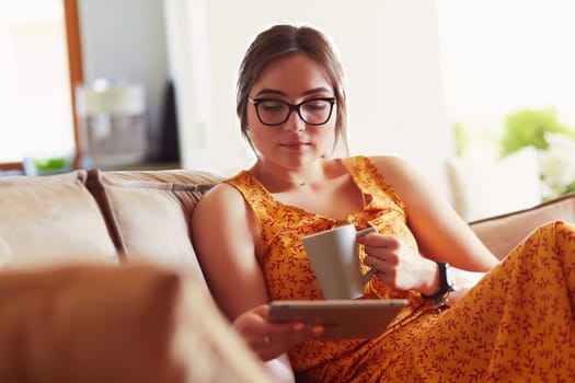Young woman uses tablet while relaxing at home with coffee on the sofa couch.