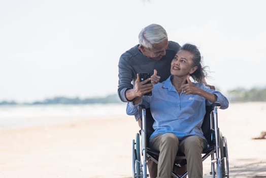 Happy Asian elderly woman sitting in wheelchair and husband is a wheelchair user smartphone taking selfie on the beach, summer vacation, Retirement couple concept