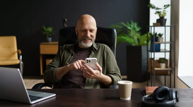 Bald and respectable man with gray beard. He captured in moment of joy, smiles while reading messages on phone. Computer, cup of coffee, and audio headphones are present on the table. High quality photo